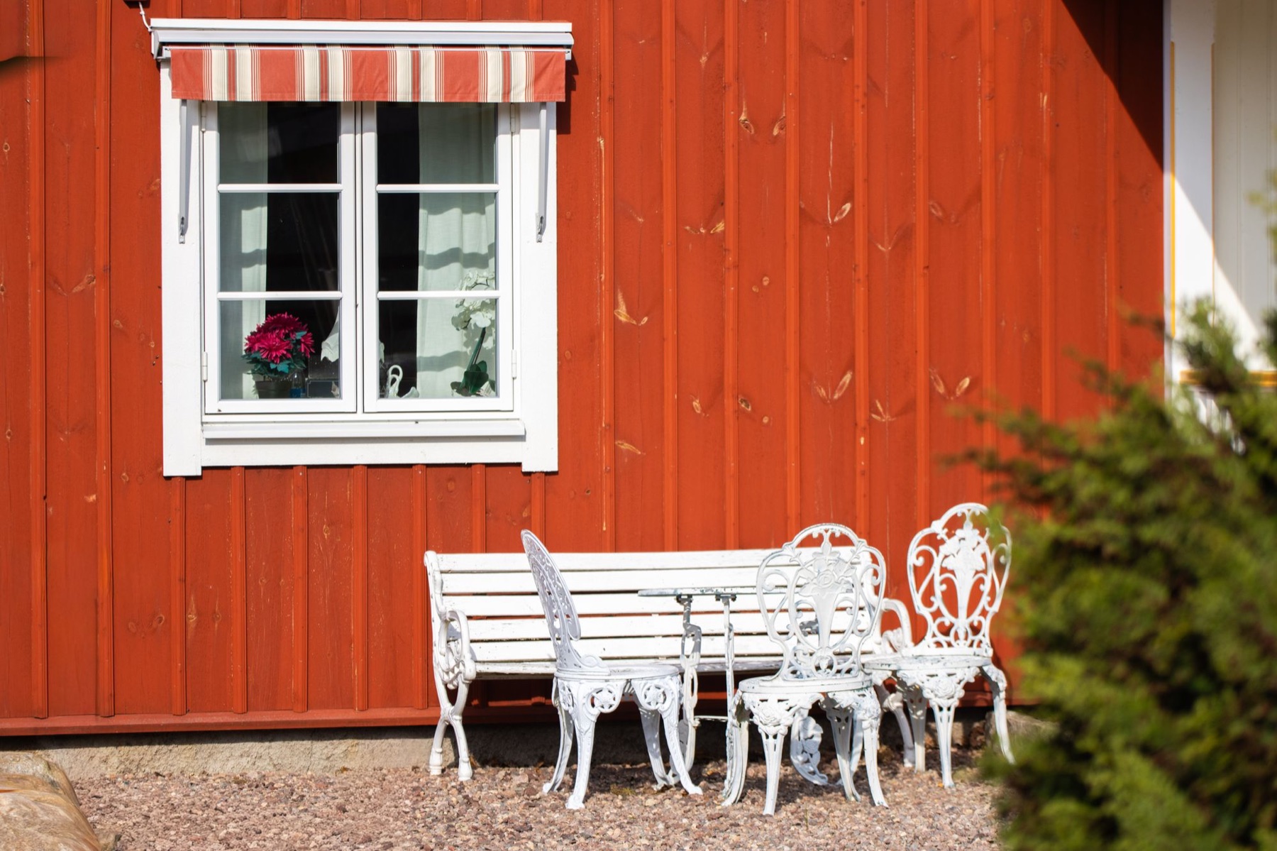 Röd träfasad med vitt spröjsat fönster, markis och trädgårdsmöbler.